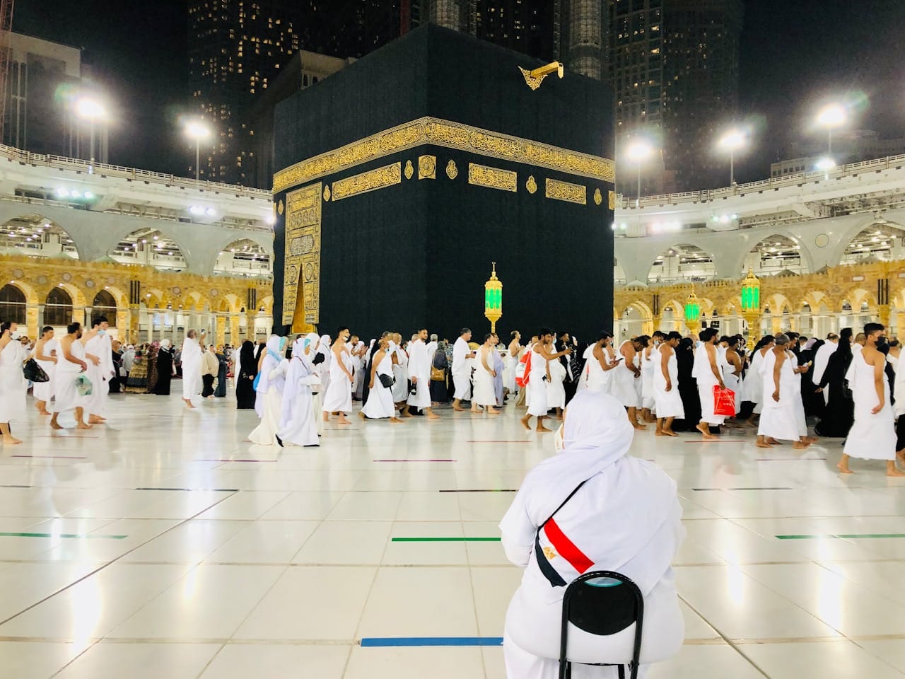 Muslim pilgrims perform Tawaf around the Kaaba in Mecca under bright lights at night.